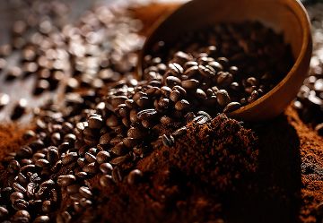 Grãos de café escorrem de uma colher de madeira sobre café moído em fundo escuro